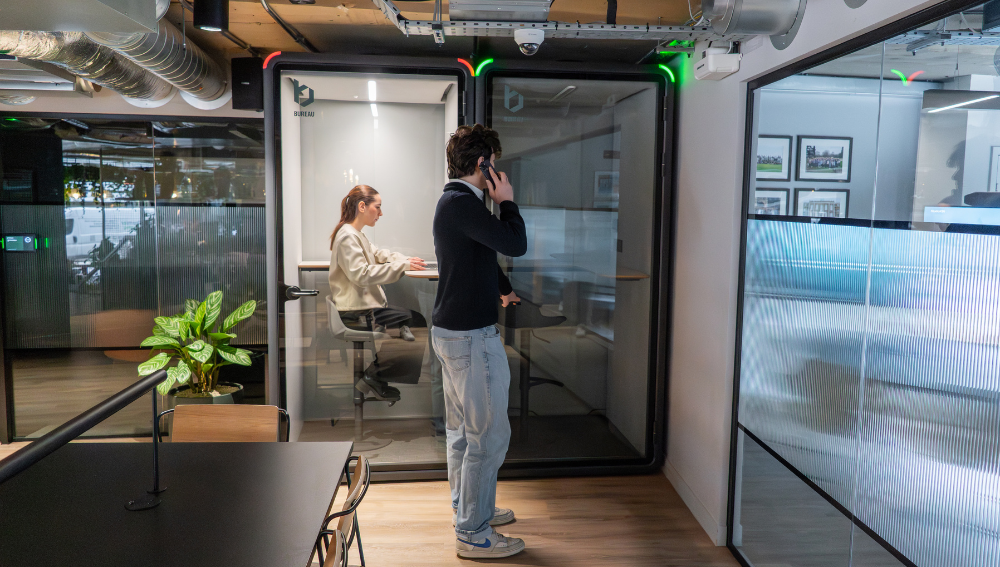 Two modern soundproof office booths in an open office environment, featuring one person working on a laptop inside and another person taking a private phone call.